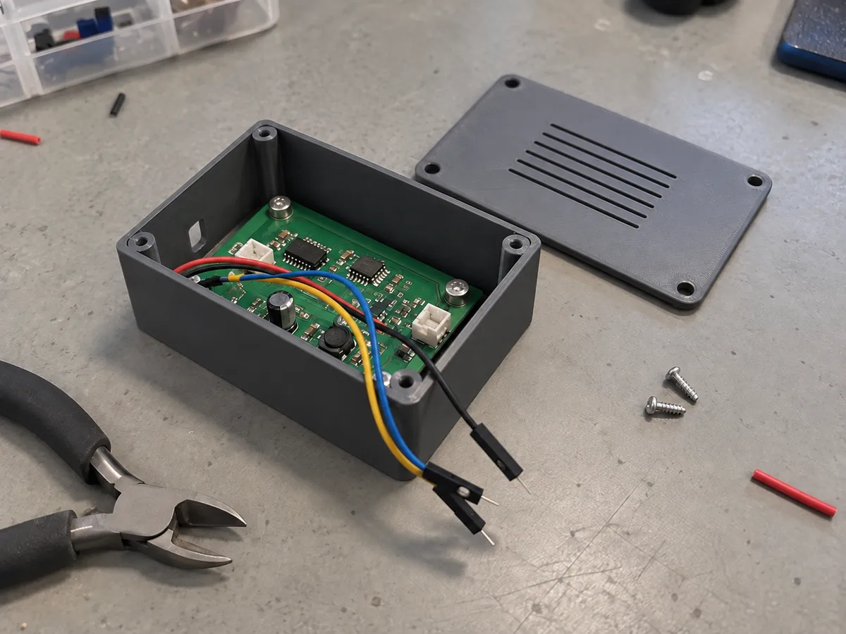 An open grey printed electronics enclosure with a green circuit board, loose jumper wires and a vented lid on a bench