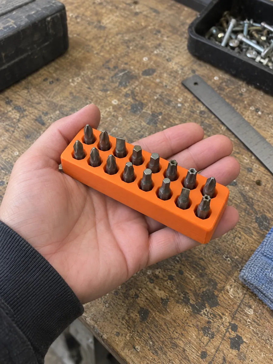 A hand holds an orange printed holder with two rows of eight metal screwdriver bits above a workbench