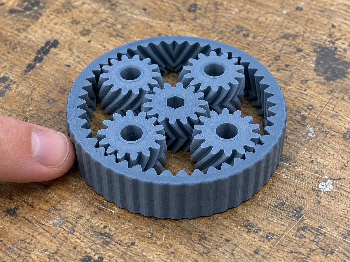 A blue printed gear bearing with a central hex drive and four surrounding gears rests beside a fingertip